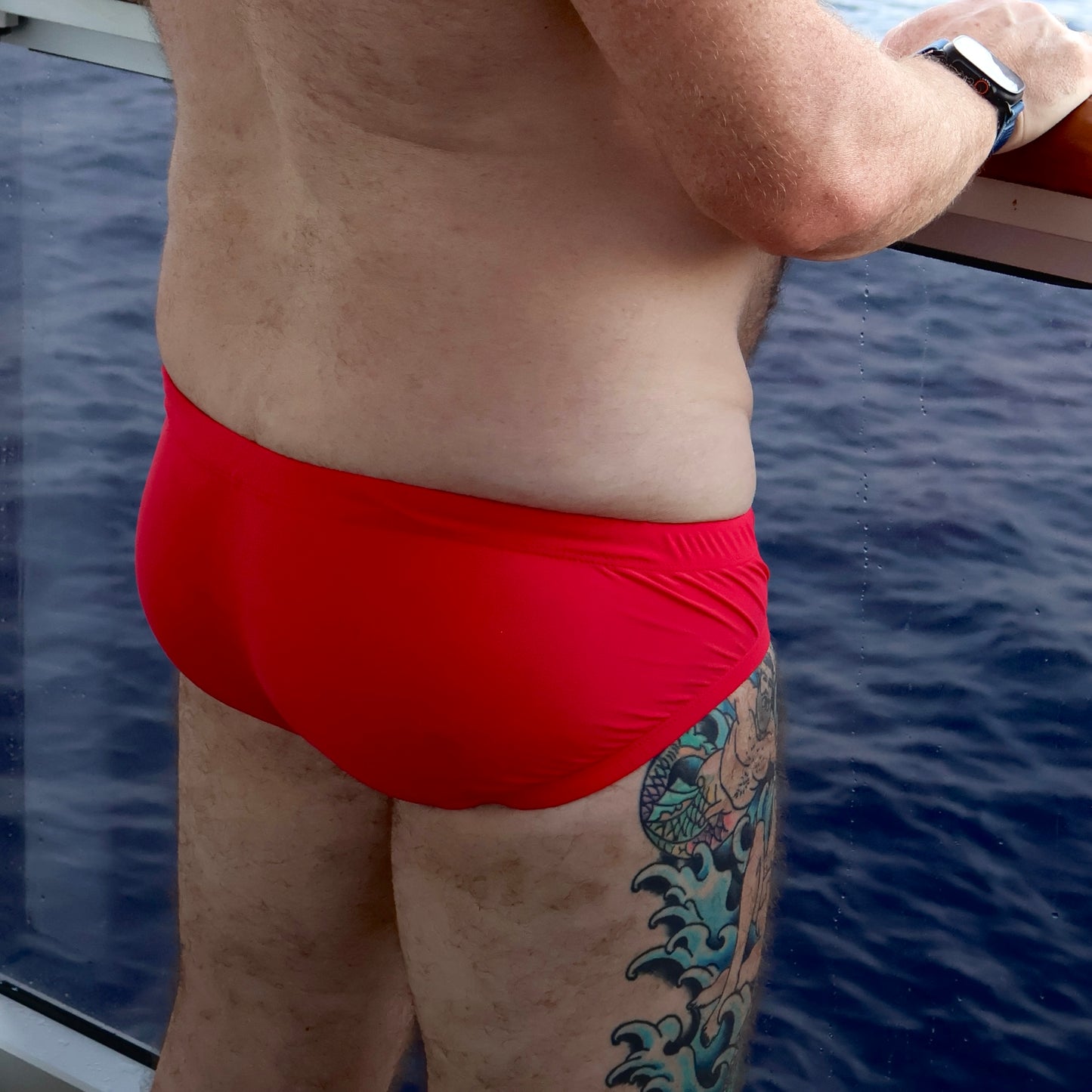 Person wearing red swim trunks with a tattoo on their leg, standing by water.