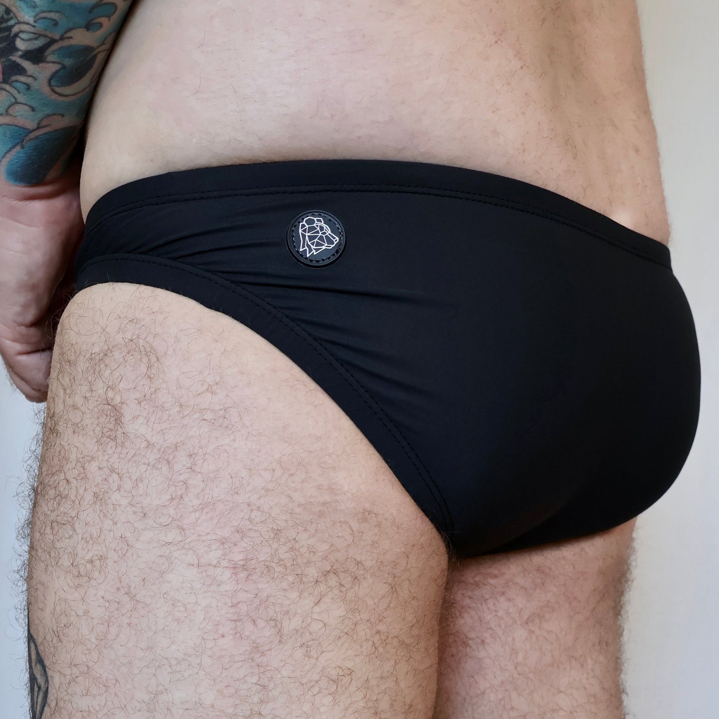 Person wearing black swim briefs with a brand logo on a neutral background