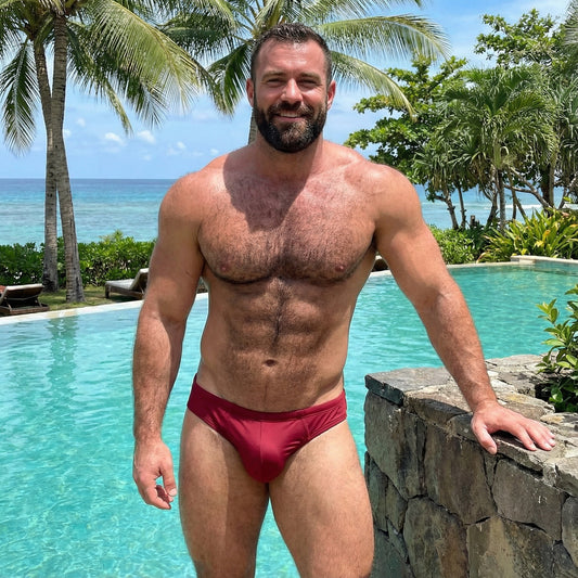 Man in red swim trunks standing by a pool with palm trees and ocean in the background