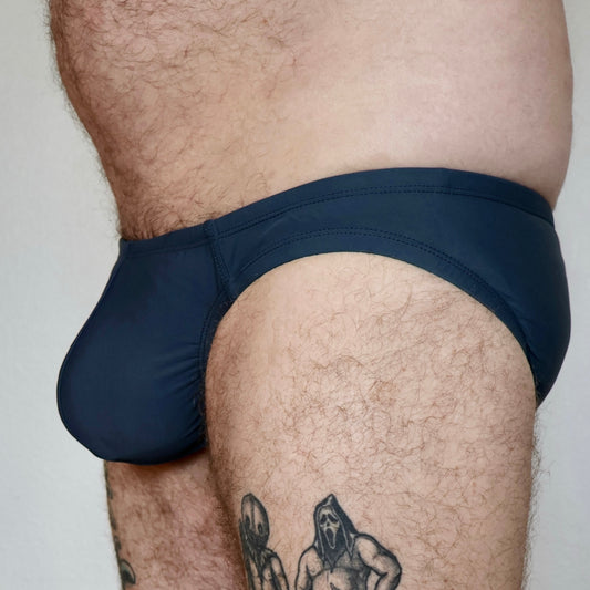 dark grey men's swim brief worn by a person with tattoos on a plain background