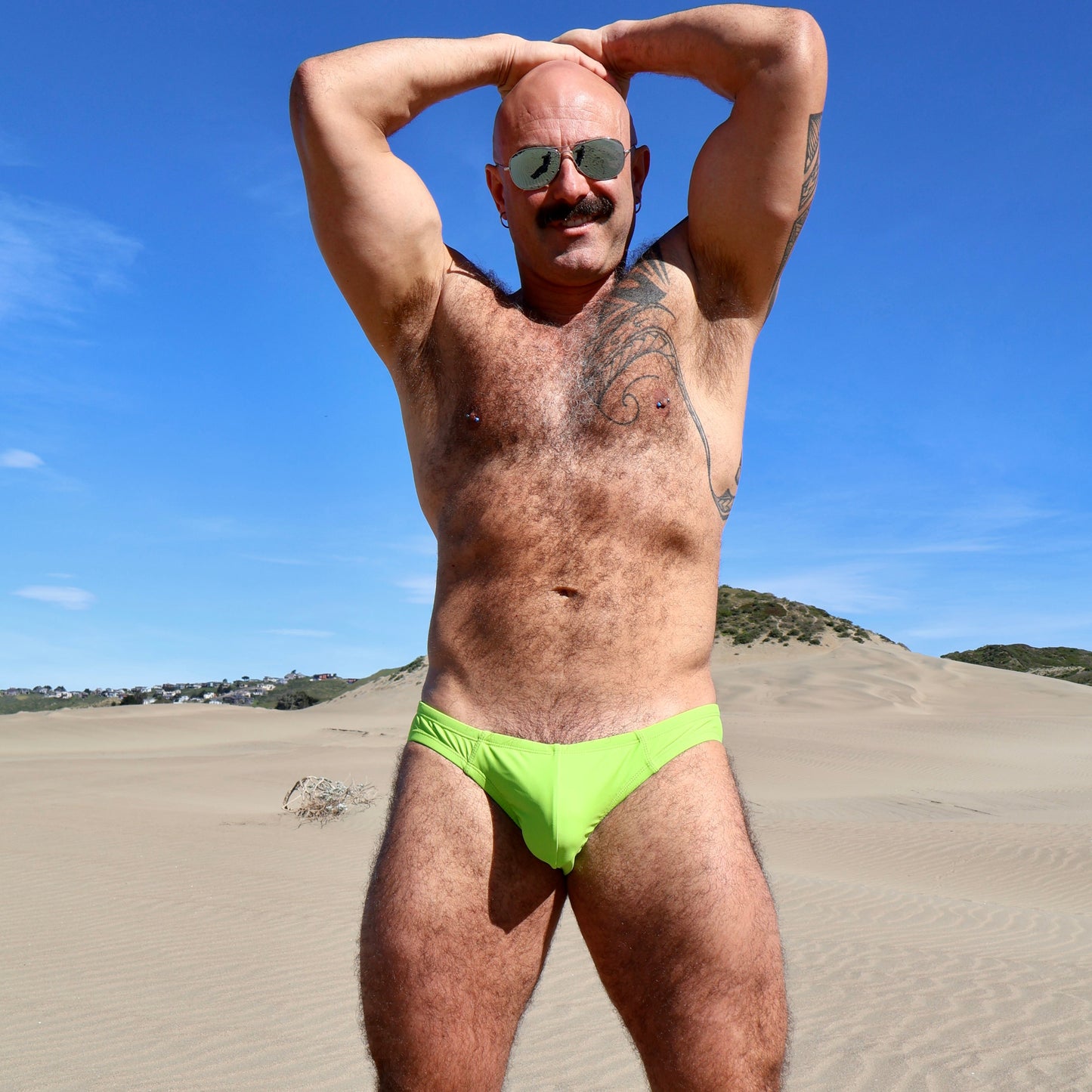 Man wearing green swim briefs standing on a sandy beach with a clear blue sky.