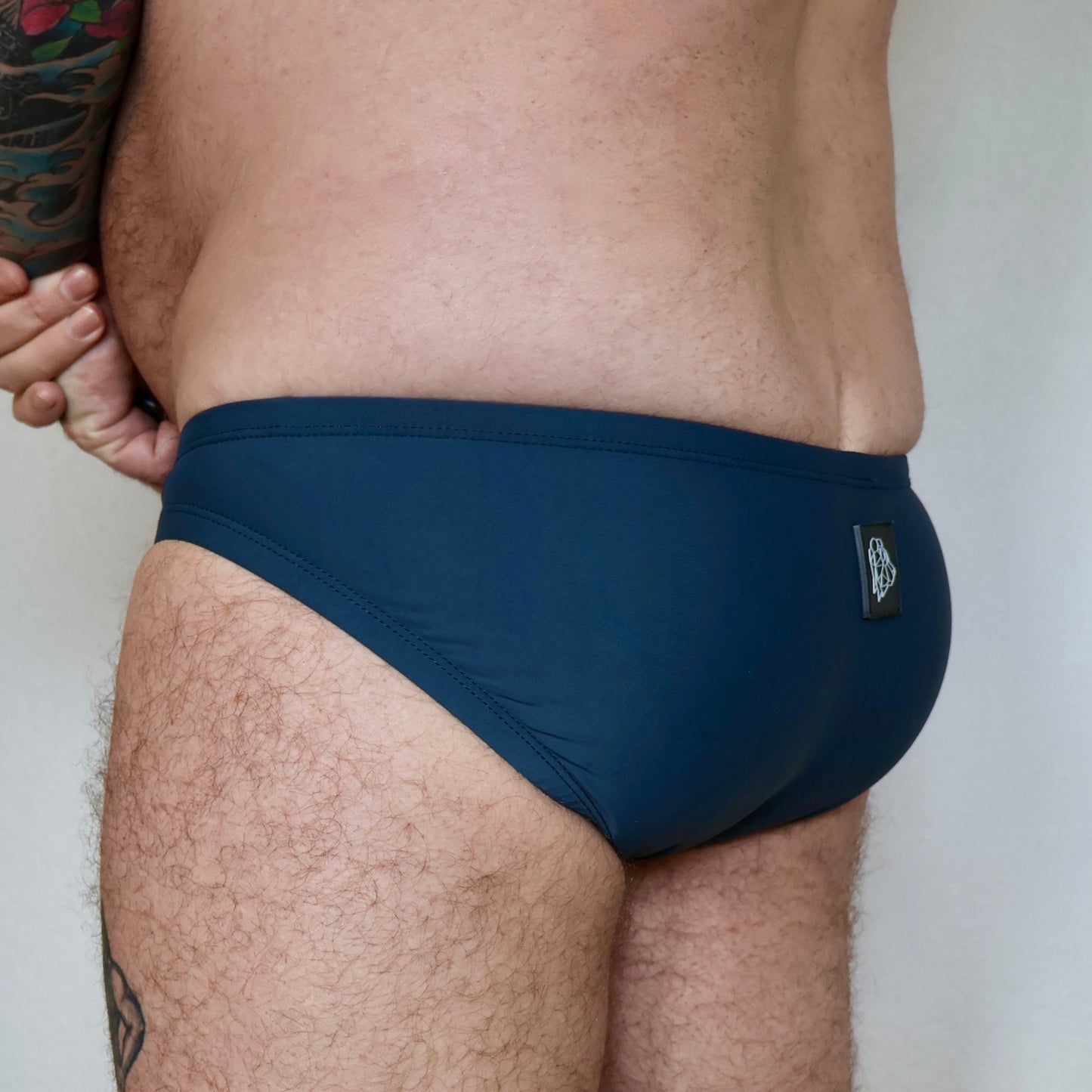 Navy blue swim briefs worn by a person with tattoos on a plain background