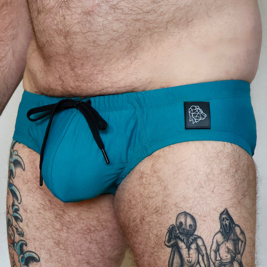 Teal swim shorts with a black drawstring and logo patch on a person with tattoos.