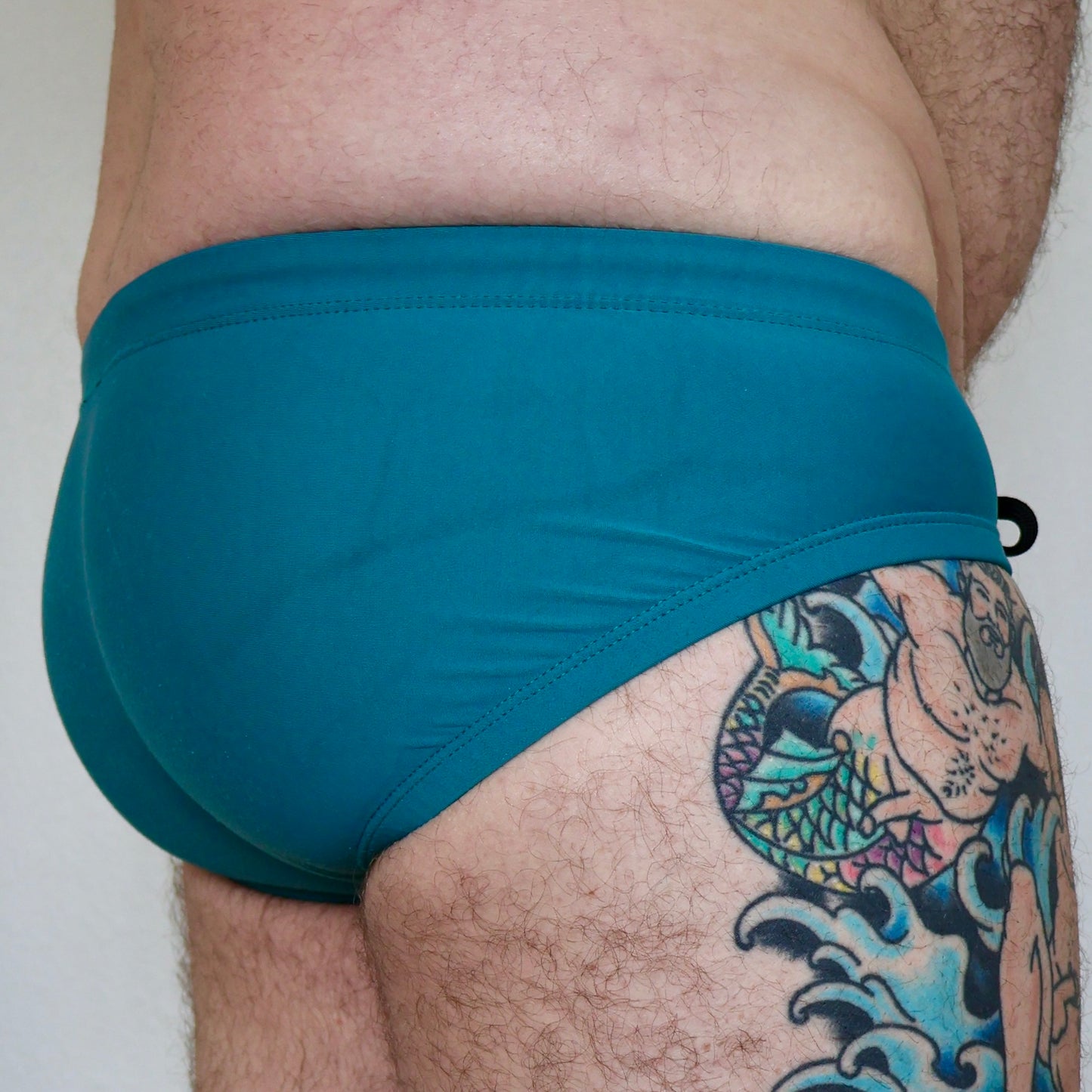 Teal swim briefs worn by a person with tattoos on a plain background