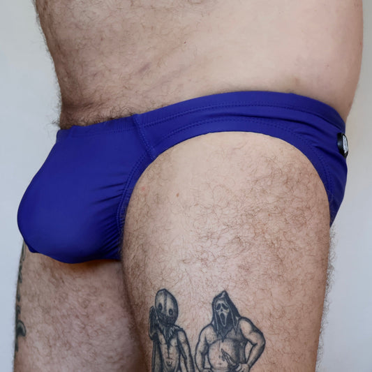 Person wearing blue swim briefs with tattoos visible on legs against a neutral background