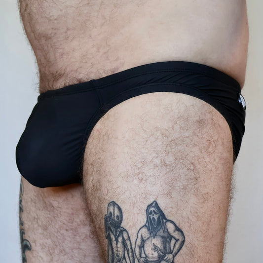 Person wearing black swim briefs with tattoos on thighs against a neutral background