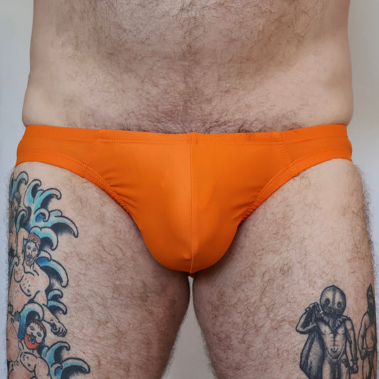 Person wearing bright orange men's swim brief with tattoos on legs against a neutral background