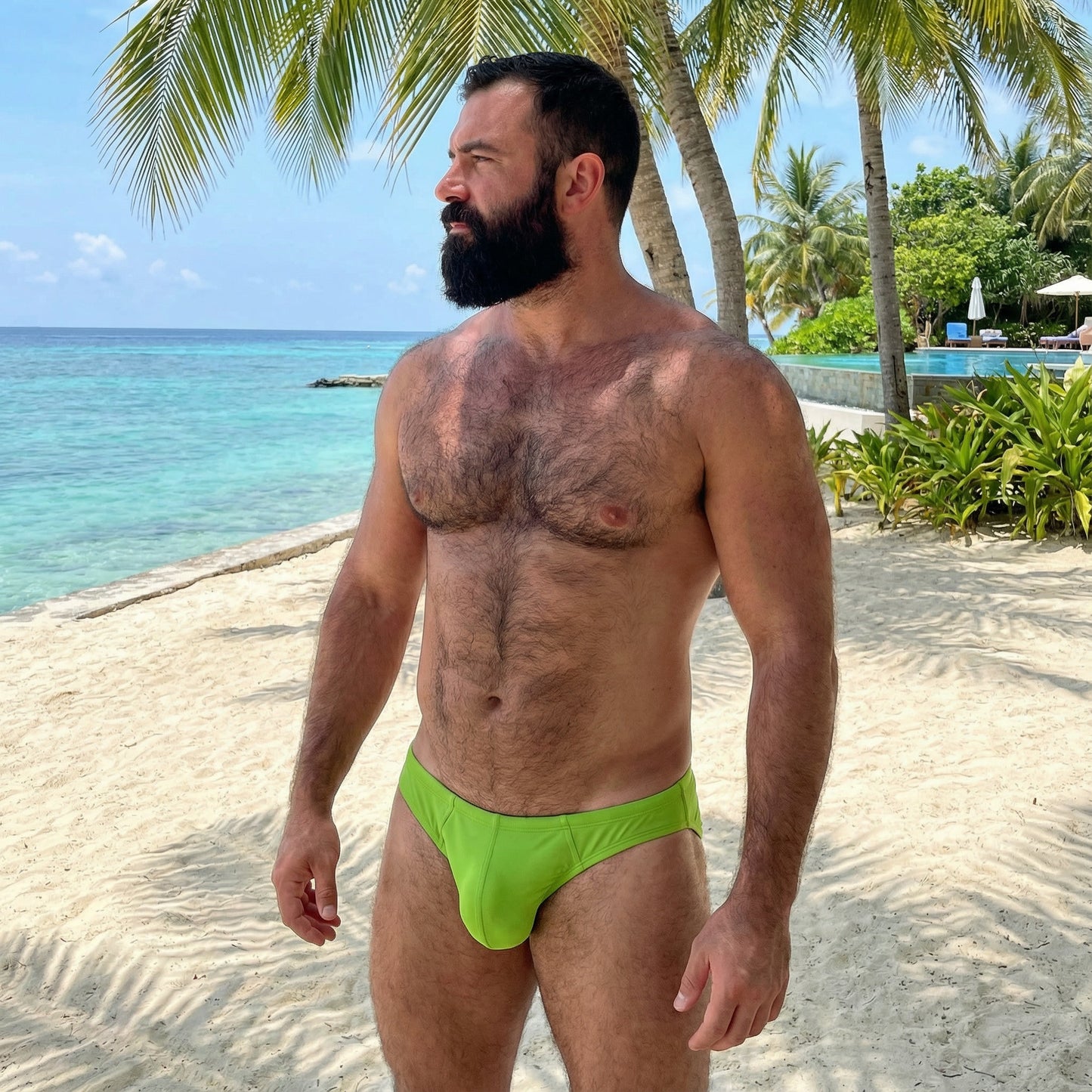 Man in green swim shorts standing on a sandy beach with palm trees and ocean in the background